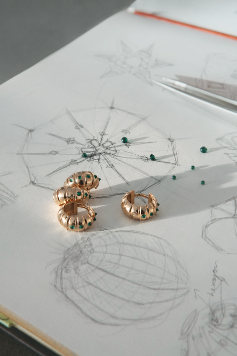 Gold huggies earrings with small green gemstone accents, displayed on a white sketchbook with concept drawings and tweezers.