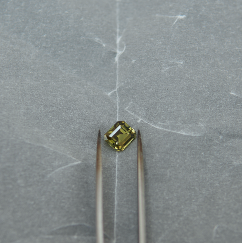 A small rectangular yellow-green gemstone held between two metal tweezers on a textured gray surface.